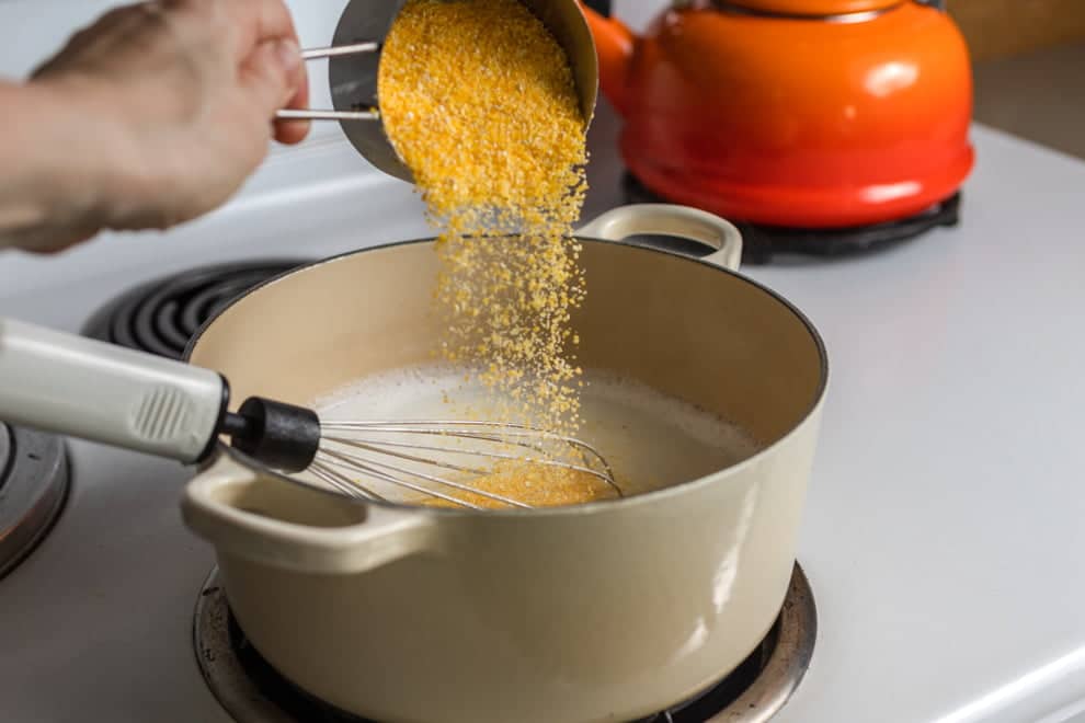 Cornmeal being poured into pot with whisk on stove.