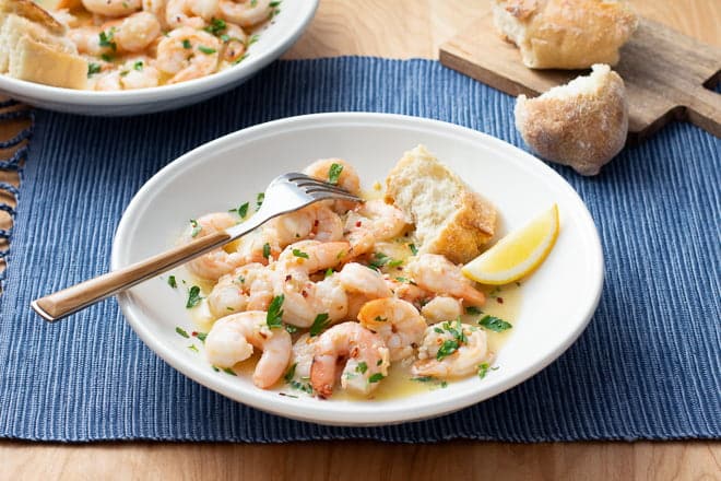 White bowl of shrimp scampi with bread and a lemon wedge.