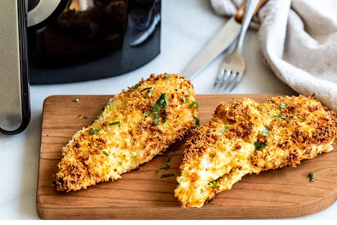Breaded chicken breasts on a cutting board in front of air fryer.