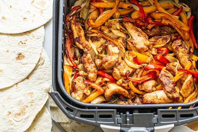 Chicken and pepper fajita mix in the air fryer basket, tortillas to the side.