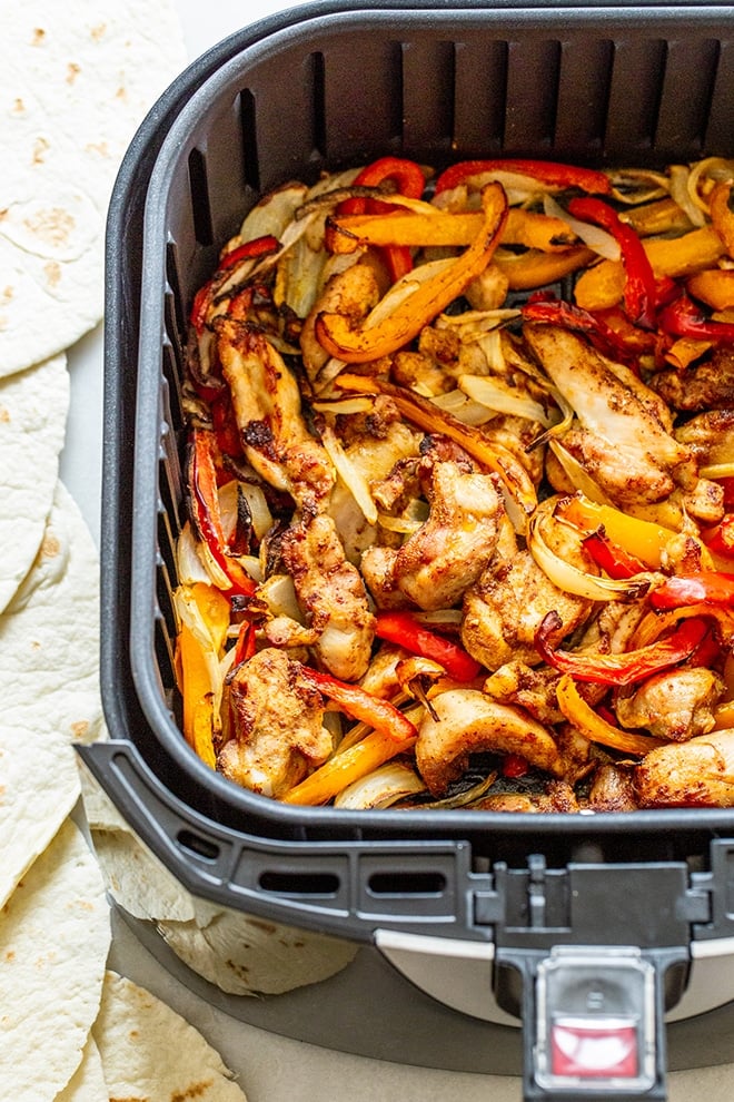 Sliced chicken, red and yellow peppers, and onions with fajita seasoning in the basket of an air fryer.