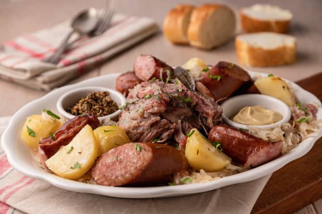 Choucroute Garnie on a large white platter. Sausages, potatoes, sauerkraut and more are included.
