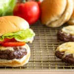 Burgers with cheese, tomatoes, and lettuce sitting on a baking sheet with rack.