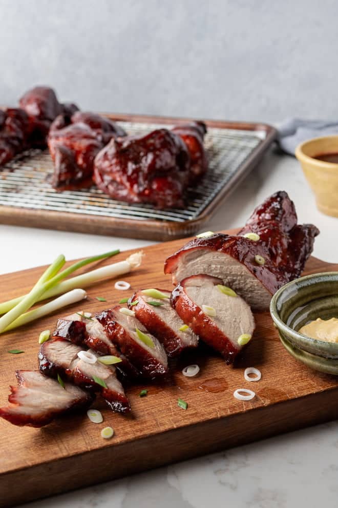 Sliced Chinese BBQ pork loin on a cutting board sprinkled with chopped green onions and mustard in a small green ceramic bowl. In the background is whole pieces of Chinese BBQ pork on a wire rack on a rimmed baking sheet.