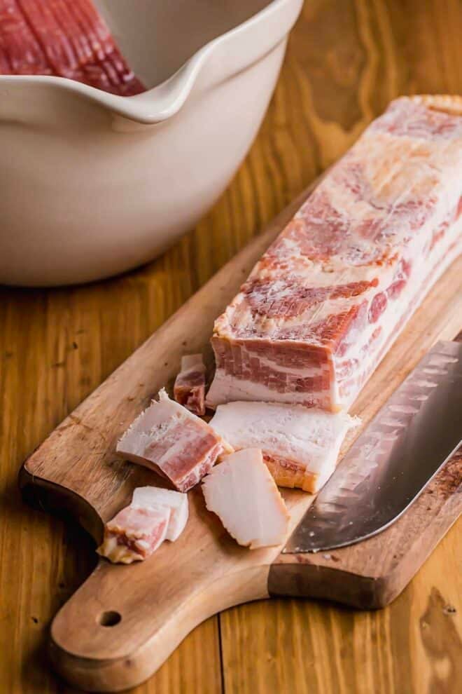 Raw bacon on a cutting board with a knife, cut into lardons.