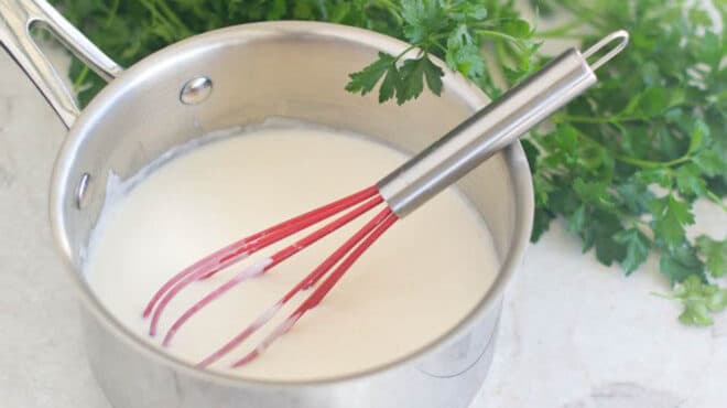White bechamel sauce in a saucepan with a red whisk.