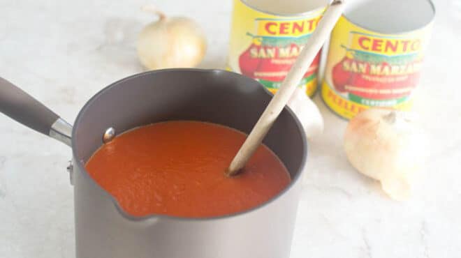 Pot of tomato sauce with wooden spoon, cans of san marzano tomatoes in background.