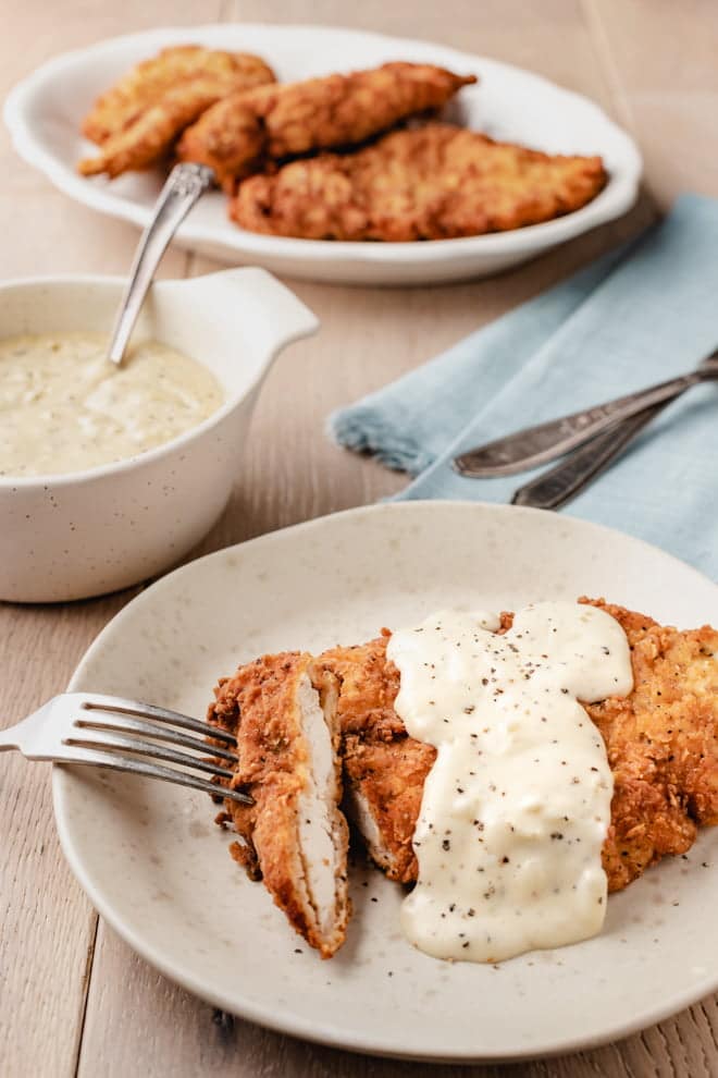 Crispy Chicken Fried Chicken smothered in a peppery cream gravy on a white plate with a fork.