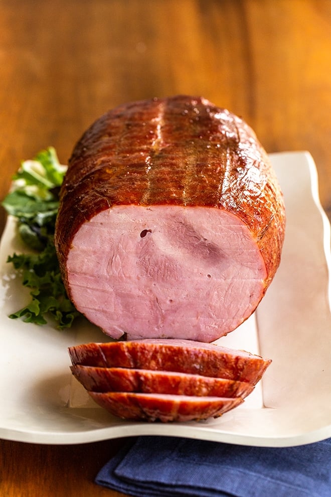 Whole, boneless ham with several slices cut on a cream rectangular platter.