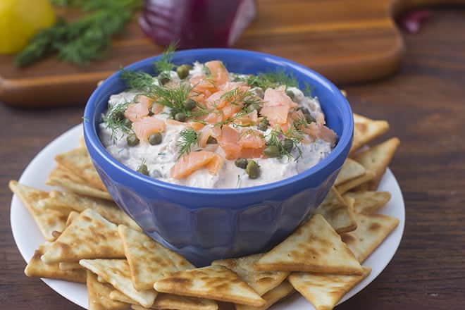 Lox Dip with capers and dill in a small blue bowl, with triangular crackers around it.
