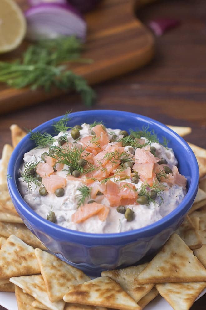 Blue bowl filled with lox dip topped with chopped smoked salmon, capers and dill fronds. The bowl is surrounded by pita chips.