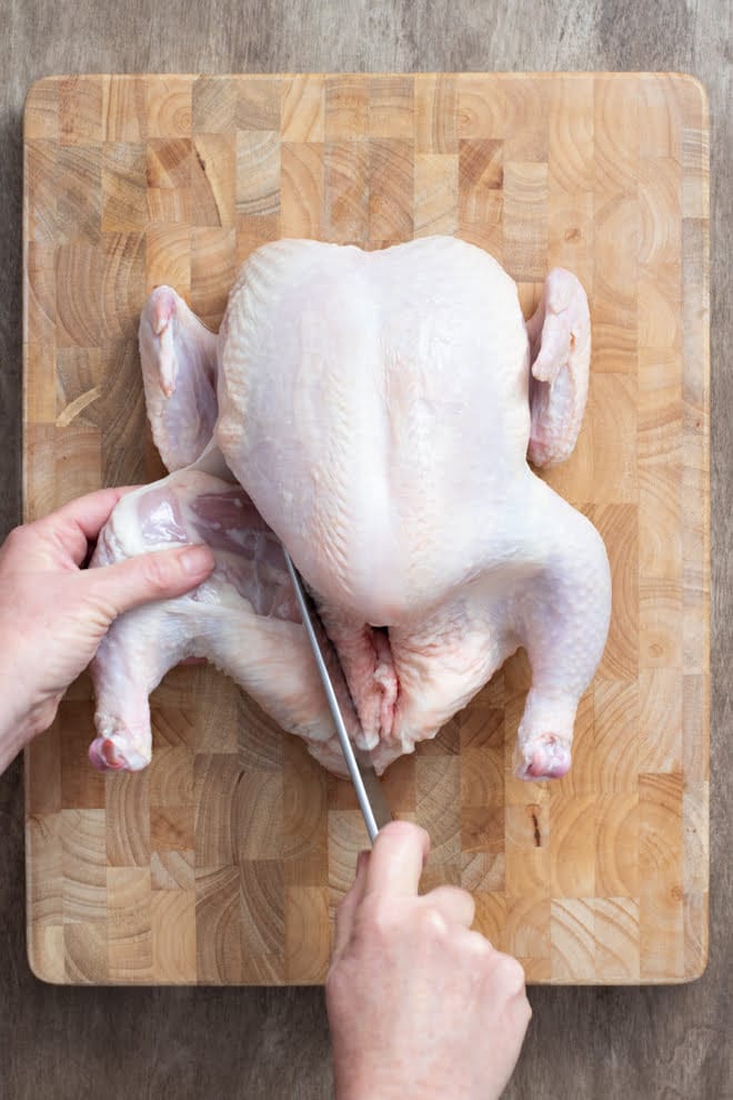 Whole raw chicken on a wooden cutting board, a hand holding onto and cutting one of the thighs.