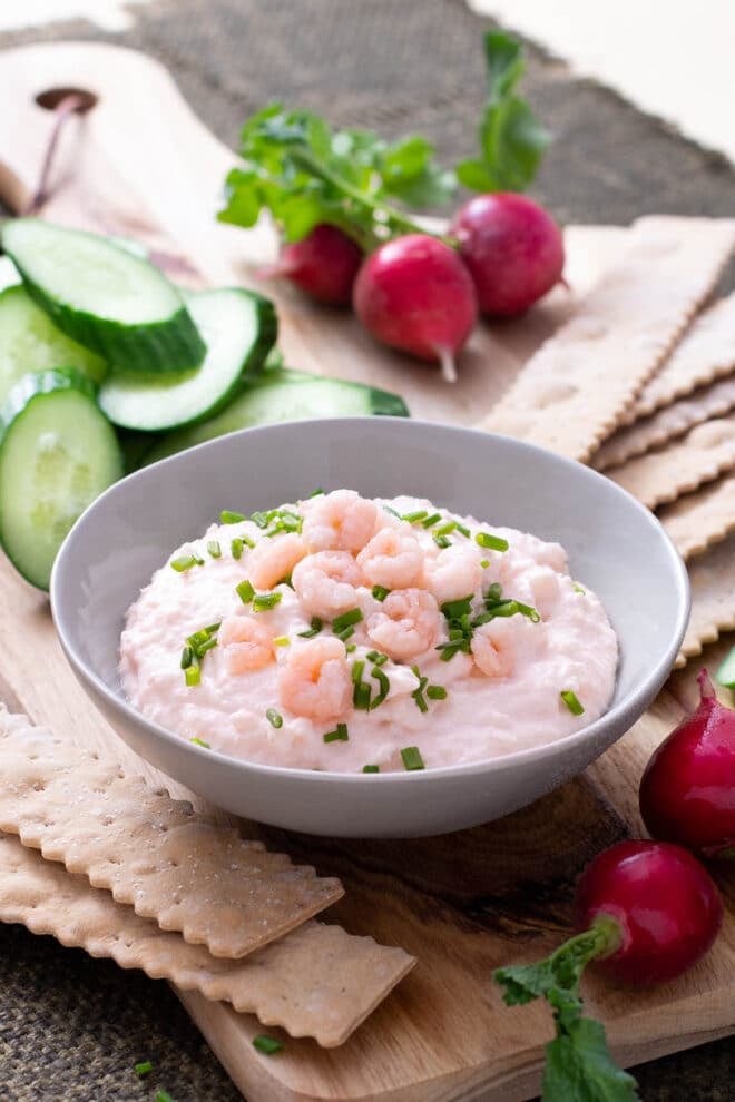 Bowl of shrimp dip with tiny shrimp and chives on top. Crackers and veggie around.