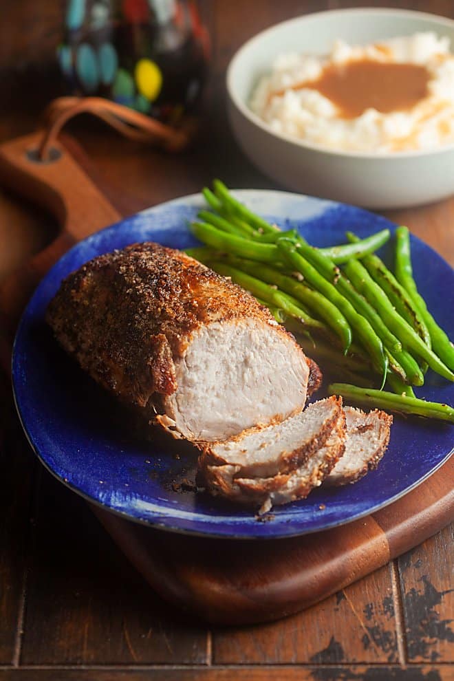 Pork loin roast on a blue plate with green beans, mashed potatoes and gravy in background.