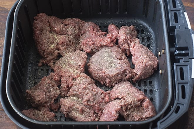 Raw ground beef in air fryer basket, seasoned with salt and pepper.