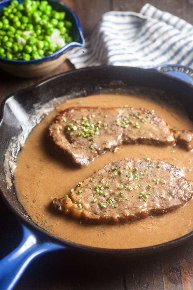 Steak and gravy in a pan with fresh herbs sprinkled on top, bowl of peas in background.