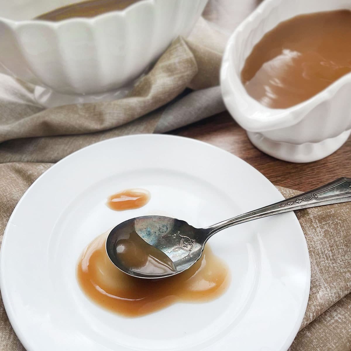 A plate with a smear of light brown gravy on it, a spoon with some gravy on it rests on the plate. A gravy boat with gravy in it is in the background