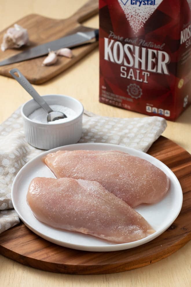 Raw chicken breasts with salt on a white plate, bowl of salt and salt box in background.