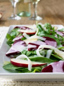 A rectangular plate with a salad of baby kale, beets, sliced apples, and Parmesan cheese shards, topped with a white creamy dressing. There are wine glasses in the background.