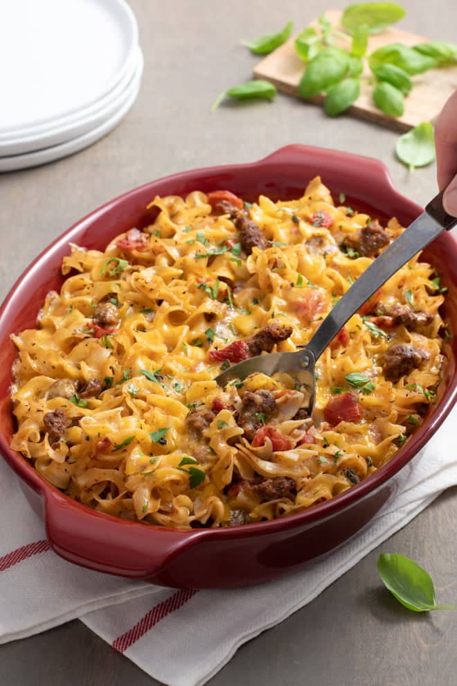 Ground Beef Casserole with noodles and tomatoes in a red oval casserole dish with a serving spoon.