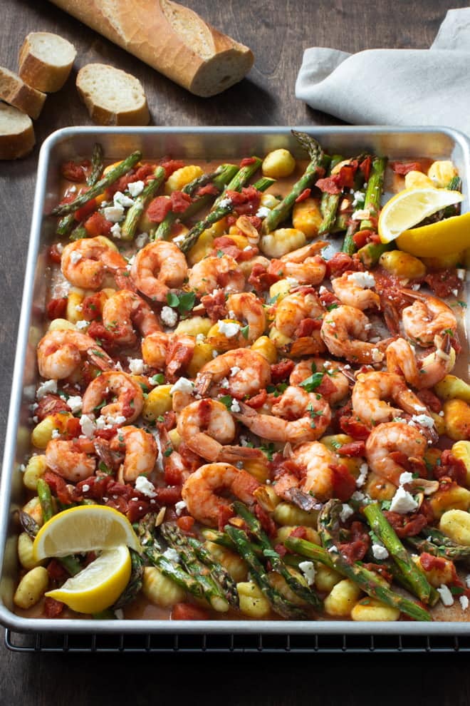 Sheet pan of shrimp, asparagus, gnocchi, and tomatoes. Lemon wedges and crumbled cheese on top.