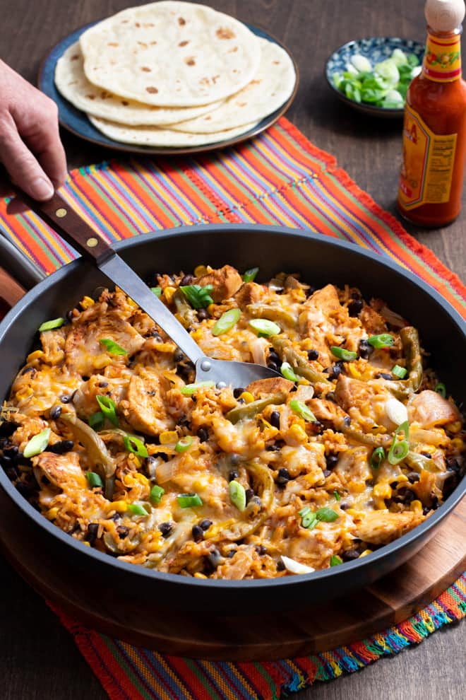 Skillet with a serving spoon going into cheesy chicken and black beans with chopped green onions scattered on top,