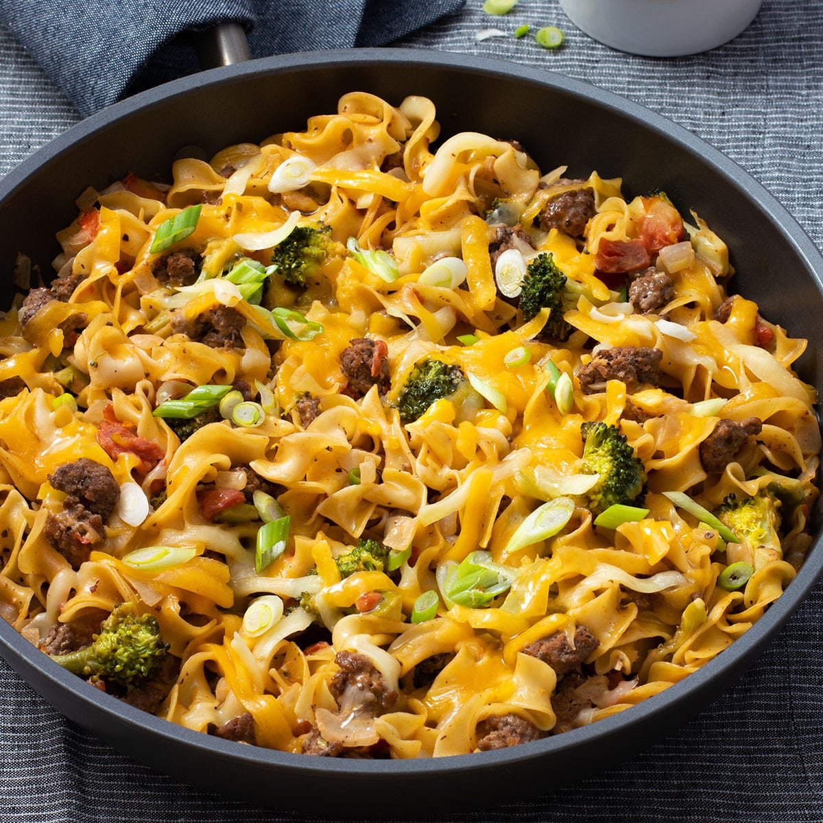 A large skillet with egg noodles, broccoli, and ground beef all cooked up and topped with cheddar cheese and green onion