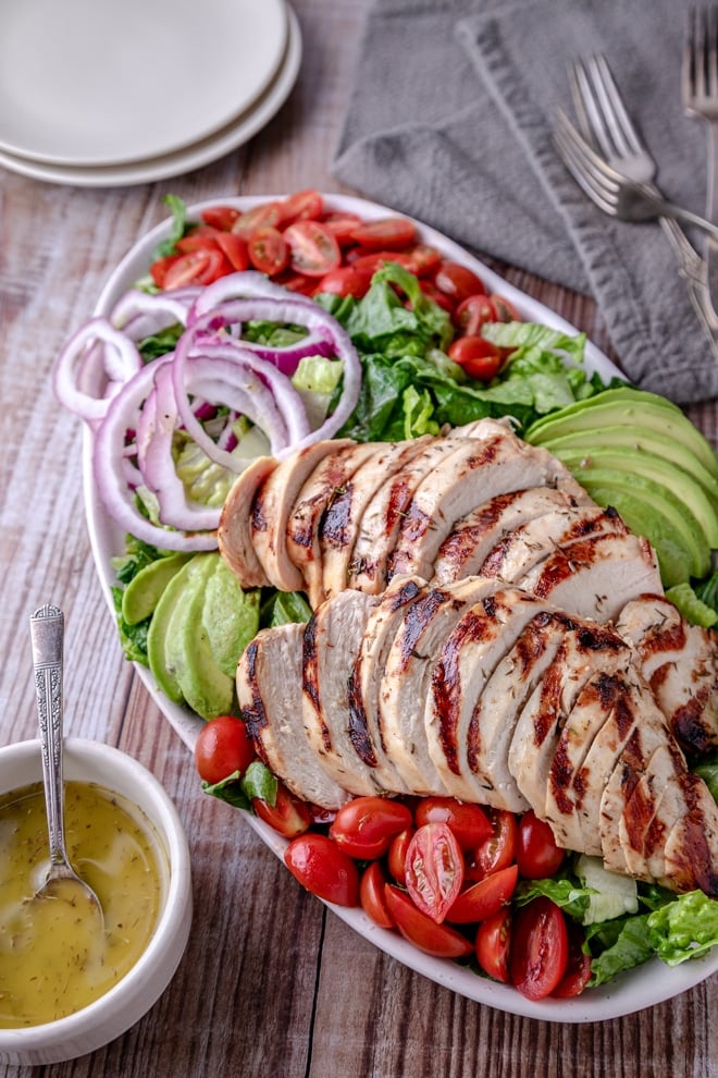 Grilled chicken breasts, sliced and on top of platter full of veggies, with a small bowl of dressing alongside.