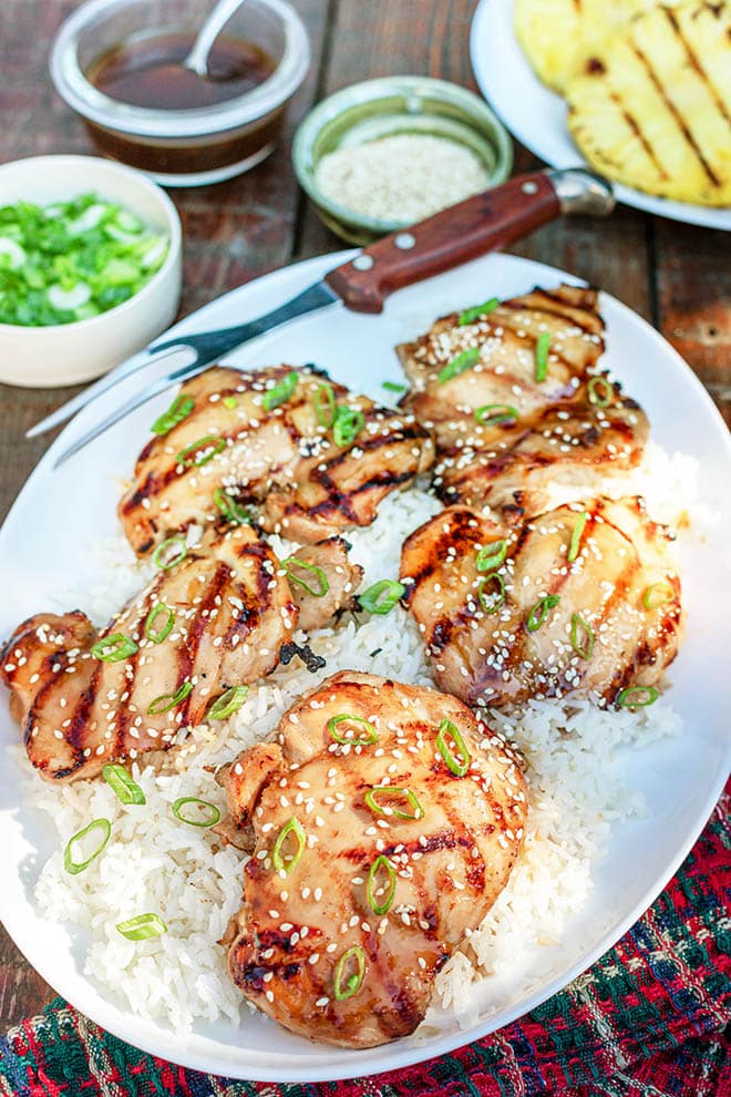 Grilled chicken thighs over white rice on a serving platter, garnished with green onion and sesame seeds. Bowl of teriyaki sauce, grilled pineapple, and garnishes are visible in the background.
