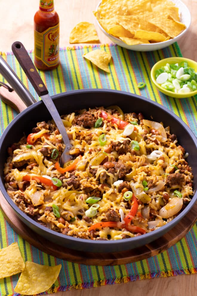 Skillet with ground beef, rice, beans, onions and peppers. Topped with melting cheese and surrounded by tortilla chips, hot sauce, and green onions.