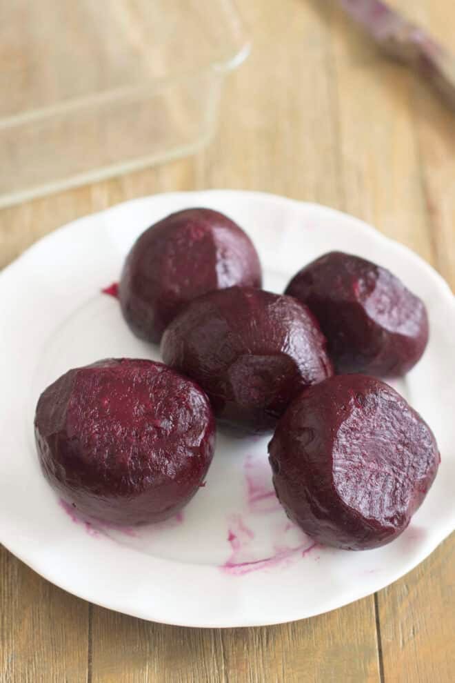 Five cooked and peeled beets on a white plate. There's smear of scarlet beet juice that has rubbed from a beet onto the plate.