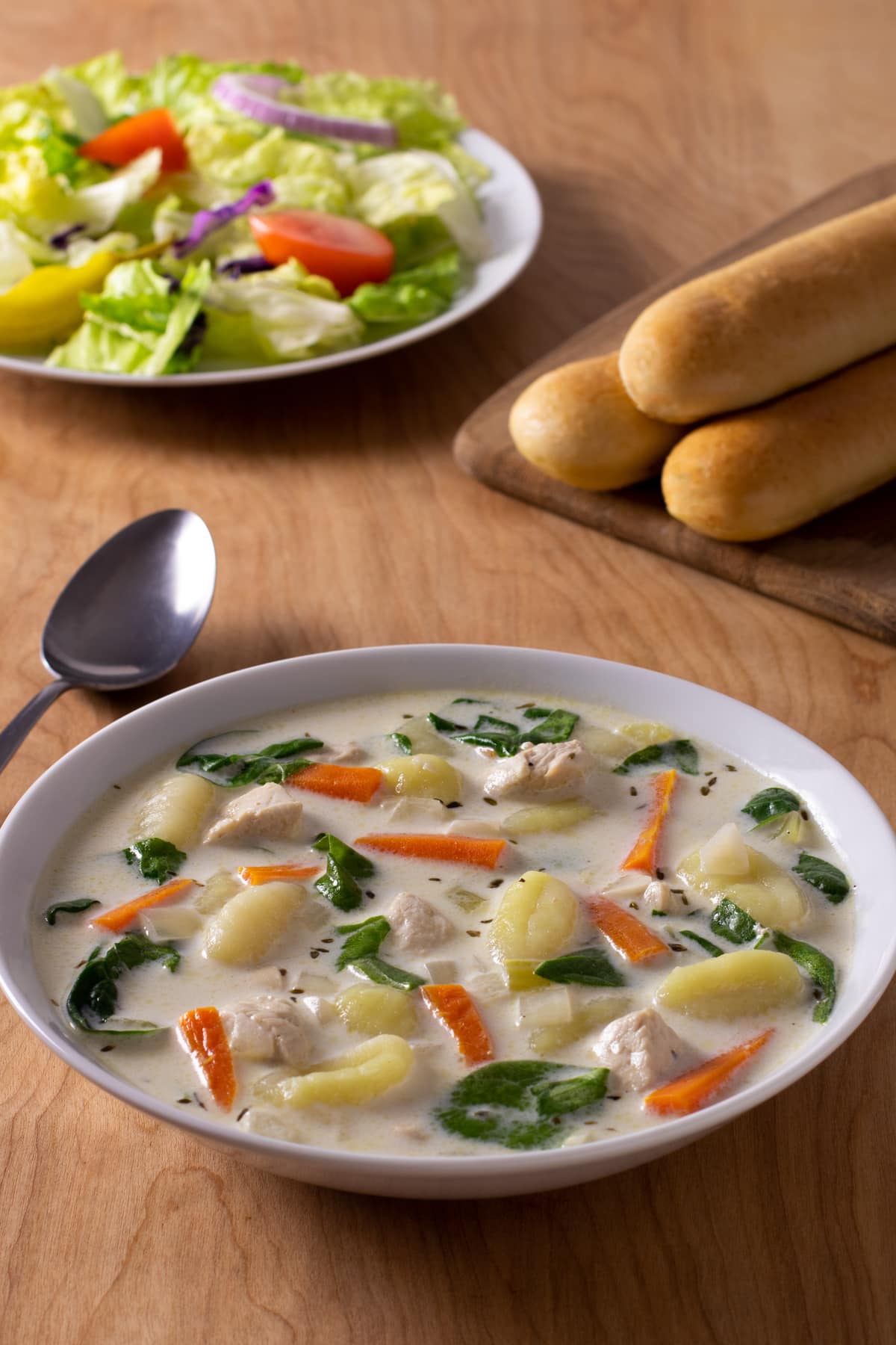 Chicken Gnocchi Soup with spinach and carrots in a creamy broth. Breadsticks and garden salad in background.
