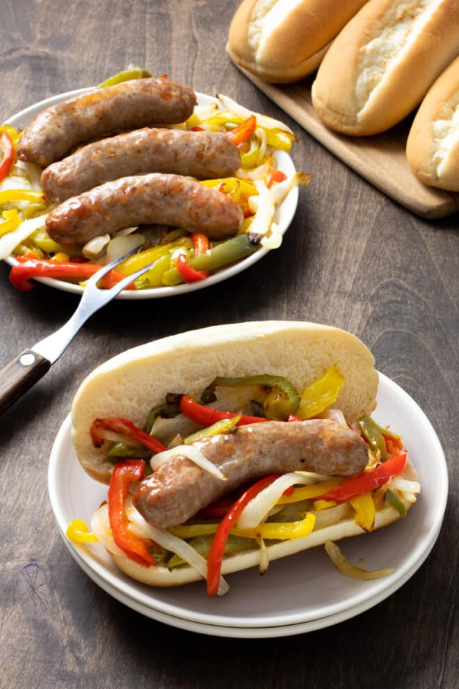 Hoagie roll with sausage, peppers, and onions on a white plate. More on a plate in background.