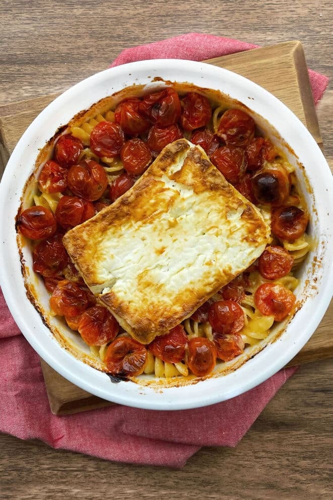 Round baking dish with cooked pasta and cherry tomatoes and browned block of feta on top.