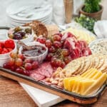 Charcuterie tray with meats, cheeses, red grapes, crackers, and white bowls of dips and olives.