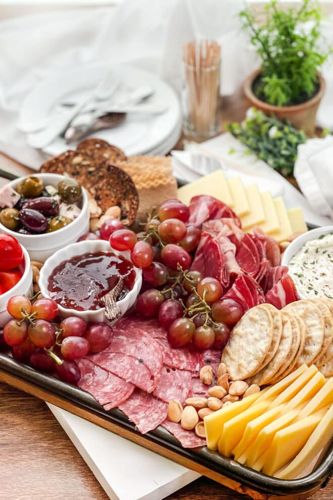 Charcuterie tray with meats, cheeses, red grapes, crackers, and white bowls of dips and olives.