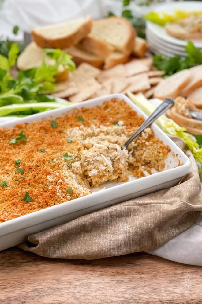 Baked Crab Cake Dip in a white dish with spoon. Celery, bread, and crackers in background.