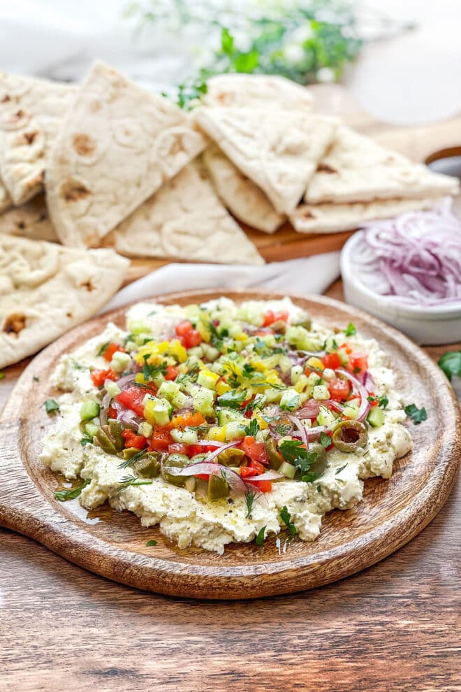 Round wooden board with whipped feta topped with onions, peppers, olives and more. Pita wedges in background.