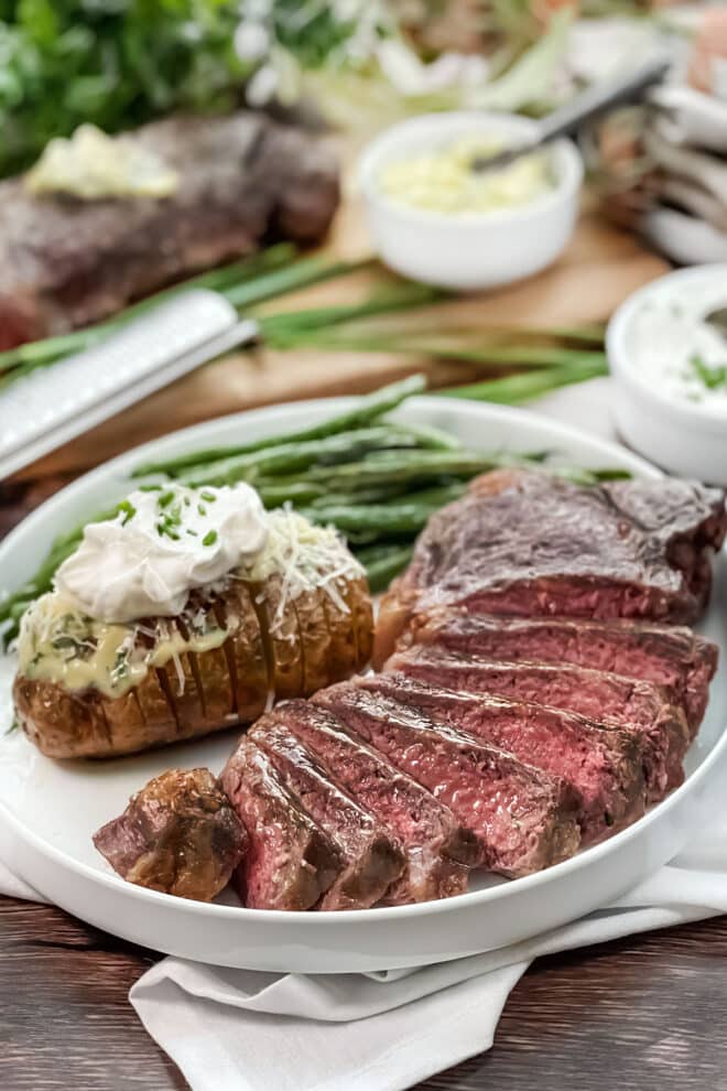Sliced steak, hasselback potato with butter and sour cream, and green beans on a white plate.