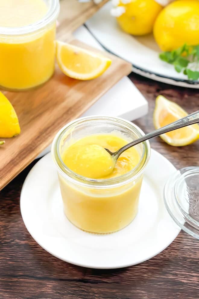 glass jar of lemon curd with spoon, lemon wedges in background