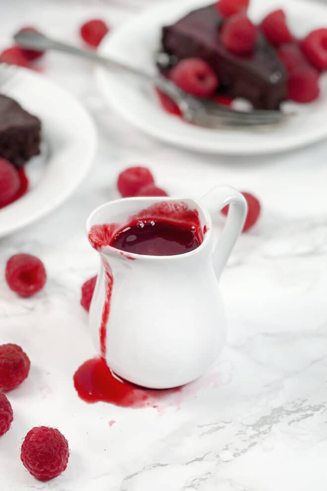 Raspberry sauce in a small white pitcher. Fresh raspberries and chocolate cake in background.