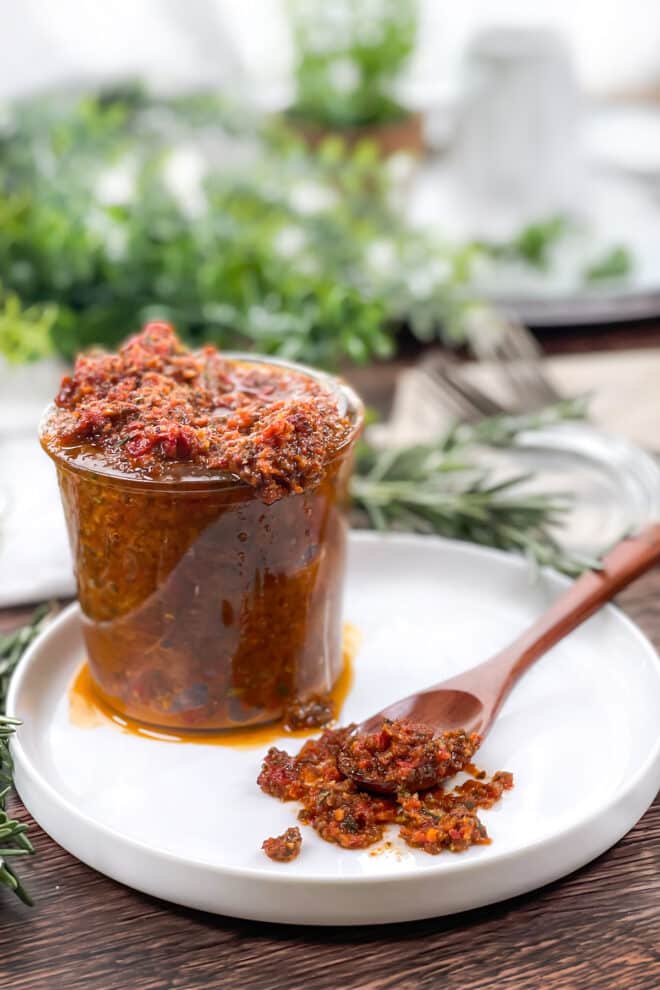 Glass jar and spoon of sun-dried tomato tapenade on a white plate.