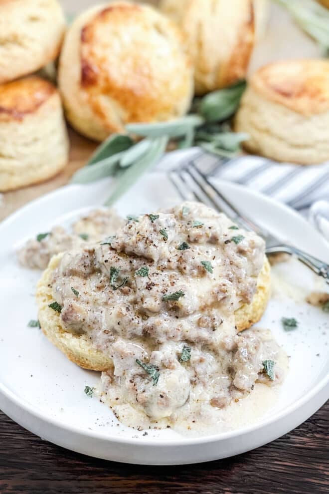 Biscuit on white plate covered with sausage gravy, more biscuits in background.