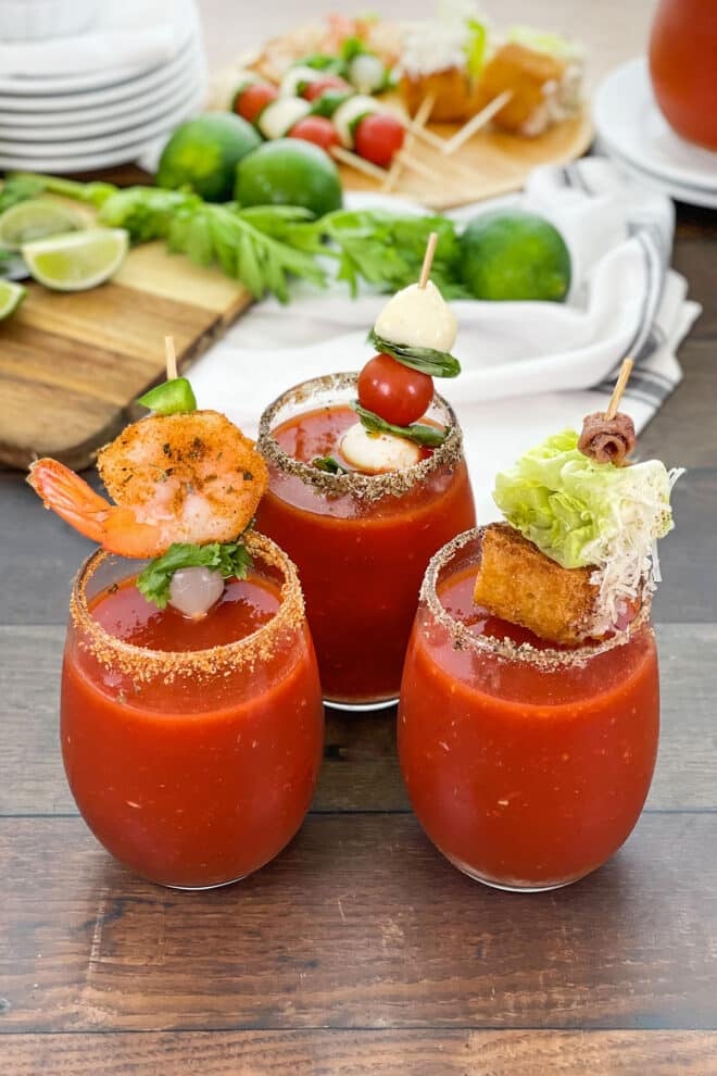 Three bloody marys in glasses, each with a skewer with different ingredients.