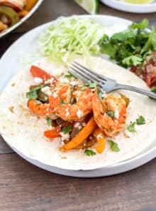 A white plate with flour tortilla on it topped with shrimp, peppers, and onions.