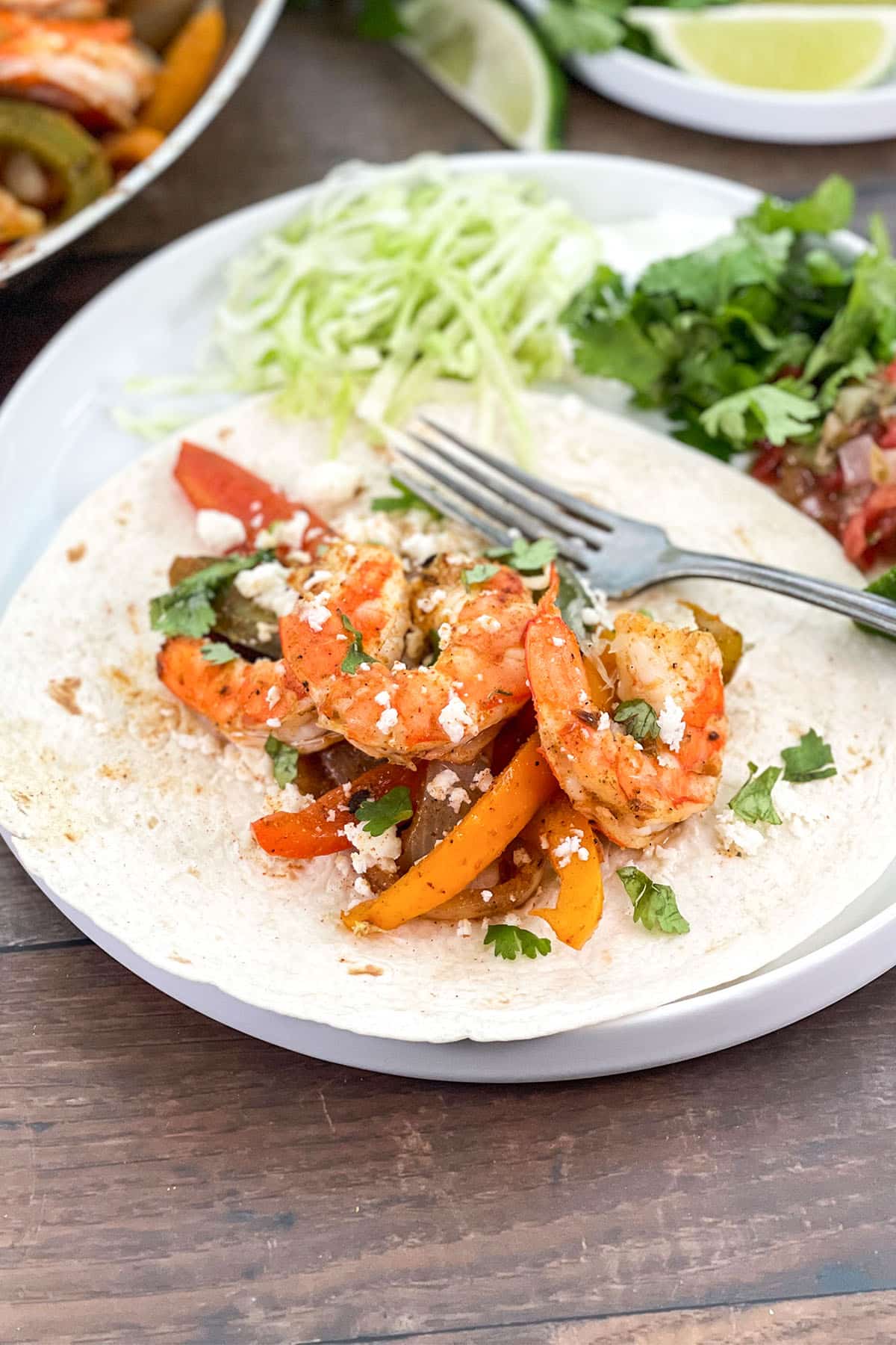 A white plate with flour tortilla on it topped with shrimp, peppers, and onions.
