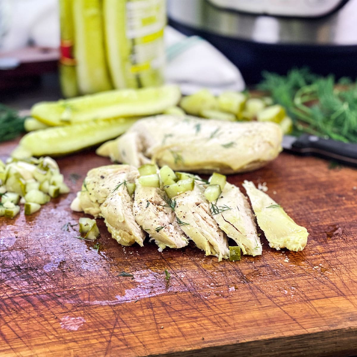 Sliced chicken breast on wooden board topped with chopped pickles, more pickles in background.