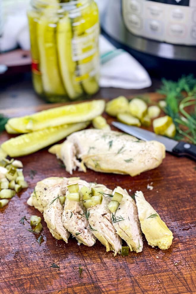 Sliced chicken breast on wooden board topped with chopped pickles and fresh dill, more pickles in background.