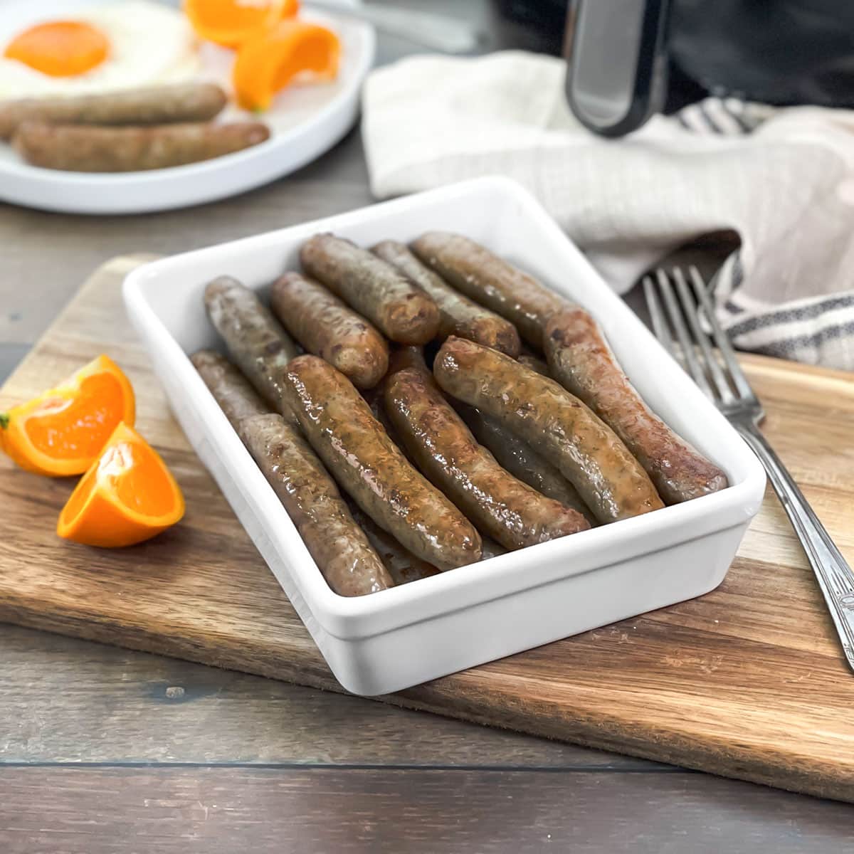 Breakfast sausage links on a white platter, with eggs and oranges in background.
