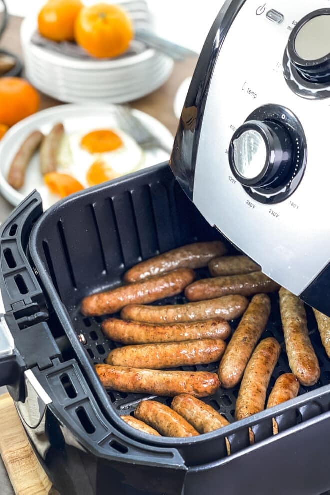 Breakfast sausage links in an air fryer basket, with eggs and oranges in background.